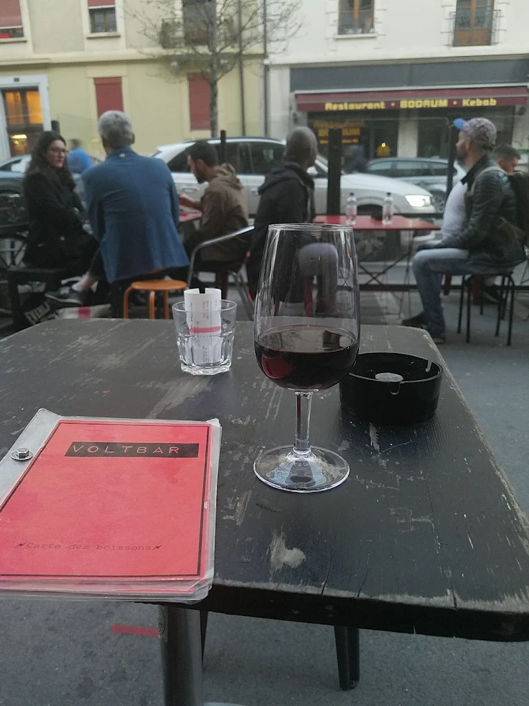 Terrasse du Volt Bar en soirée : carte des boissons et verre de vin sur une table en bois, rue de Plainpalais en arrière-plan.